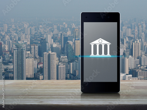 Bank flat icon on modern smart mobile phone screen on wooden table over city tower and skyscraper, Mobile banking concept