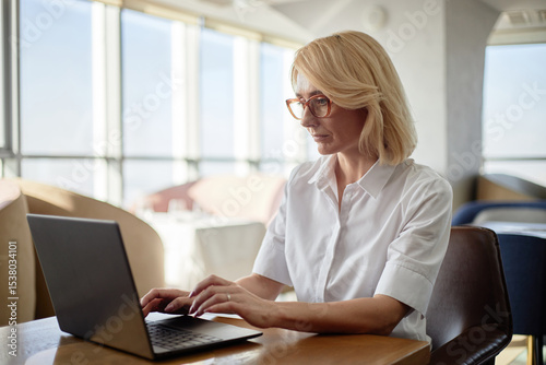 Caucasian middle aged woman working on laptop in restaurant setting, sitting at table and typing on keyboard, focused on screen, wearing glasses, natural light coming through windows Free Download Caucasian middle aged woman working on laptop in restaurant setting, sitting at table and typing on keyboard, focused on screen, wearing glasses, natural light coming through windows - GFXLoad