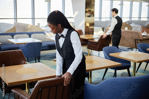 Young adult Black woman arranging chair in modern restaurant dining area while young Caucasian man in uniform working in background