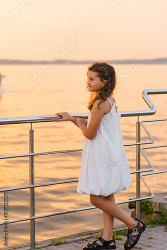 Child girl stands on pier overlooking body of water. She is wearing white dress and is smiling