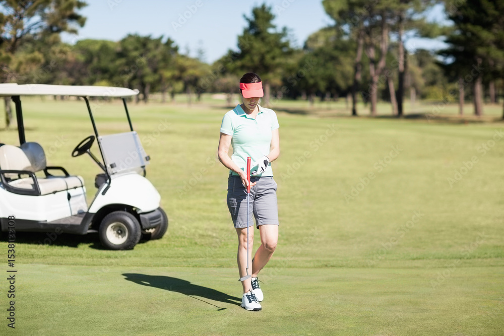 Obraz premium Female golfer standing on putting green holding putter and golf ball near white golf cart