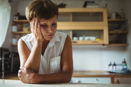 Senior woman leaning on hand at wooden kitchen counter near window, with coffee machine and mugs