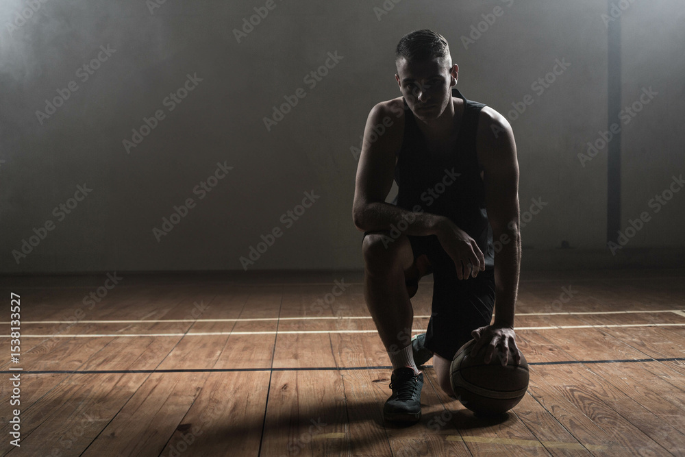 Fototapeta premium Male athlete kneeling on lined court holding basketball under lighting and smoke drifting by beam
