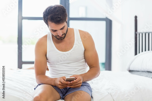 Man sitting on edge of white-linen bed using smartphone by black-framed window with green foliage