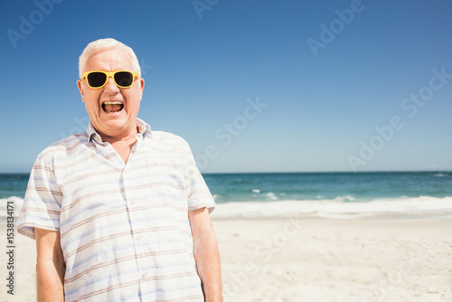Senior man standing on sandy shore at beach wearing striped shirt and yellow sunglasses, copy space