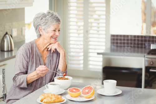 Senior woman sitting at breakfast table in modern kitchen, holding spoon near bowl of fruit cereal