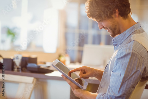 Man is interacting with white tablet on desk near coffee mug and plant under warm sunlight