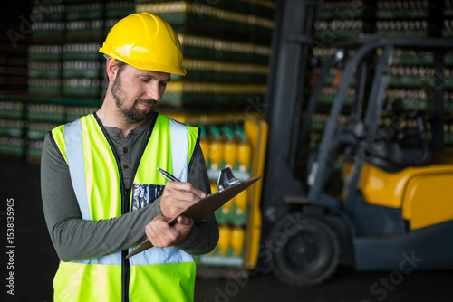 Man wearing safety vest, hardhat writing on clipboard with pen by forklift in warehouse, copy space