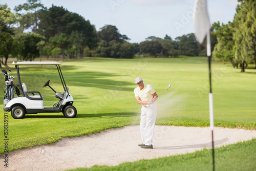 Senior man taking bunker shot hitting golf ball toward flagstick in sand trap, copy space