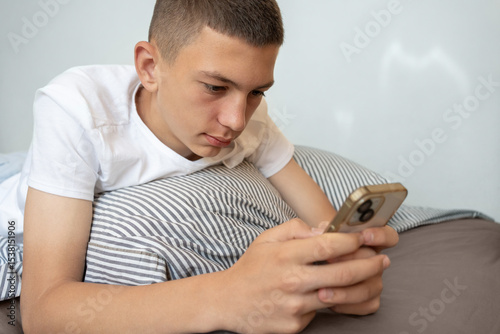 Teen boy relaxing in bed while using smartphone. Relaxed teenage boy lies in bed, looking at phone, watching videos, reading messages, or browsing social media in casual, tech-savvy moment