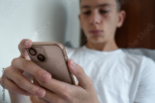 Teenage boy uses smartphone in bed. Focus on the phone in his hands, with background and face blurred, showing private digital interaction.