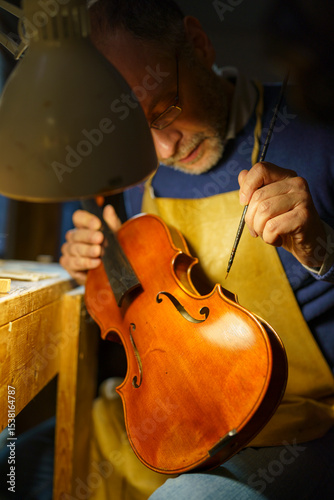 Fotografie Skilled luthier inspecting handcrafted violin in wooden workshop, warm lamplight