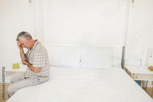 Senior man pressing forehead with hand and elbow resting on pillows on white bed near bulbs