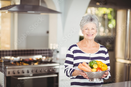 Metal colander holding fresh carrots and peppers is sitting on marble countertop in modern kitchen