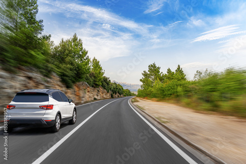 A dynamic low-angle shot captures a modern white SUV driving quickly along a winding coastal highway, conveying motion and the freedom of a scenic road trip under a bright blue sky.