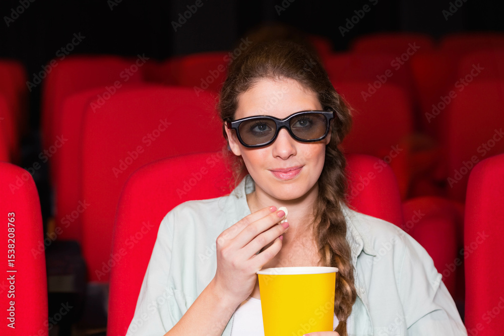 Obraz premium Teenage white female wearing black 3D glasses holding yellow popcorn bucket in darkened auditorium