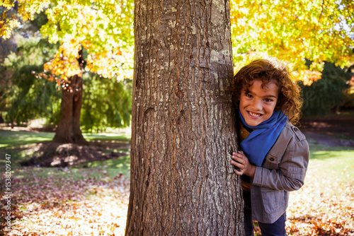 Boy peeking from behind tree trunk in park among fallen leaves wearing blue scarf, copy space