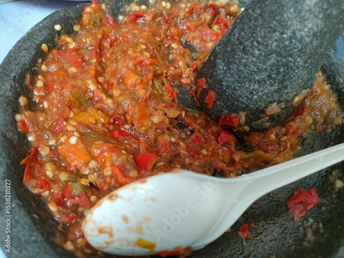 Vibrant red homemade Indonesian sambal (chili paste) being ground in a traditional stone mortar with a pestle and a white spoon, showcasing spicy culinary preparation.