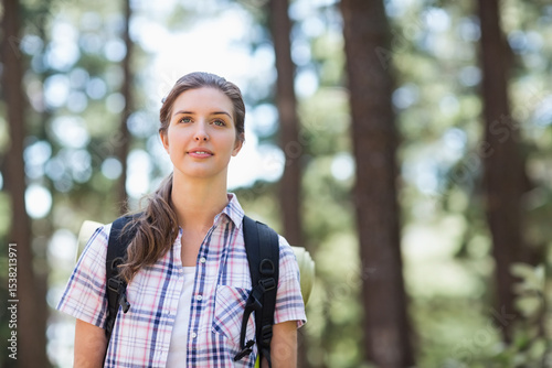 Female hiker standing in sunlit pine forest wearing plaid shirt and backpack with rolled mat