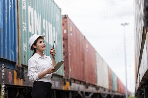 A female transport officer in a hard hat holds a clipboard and walkie-talkie, inspecting cargo containers on a freight train during export operations at a logistics and shipping terminal.