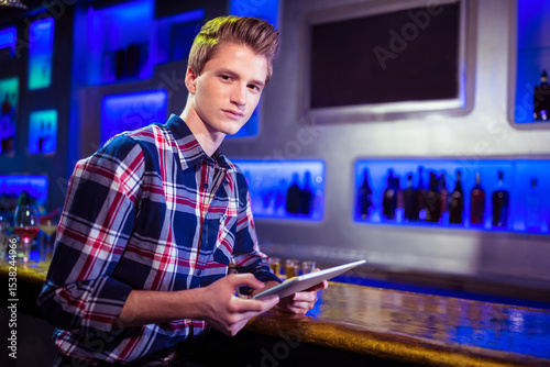 Tablet computer resting on wooden bar counter near wine glass under neon blue backlit bottles