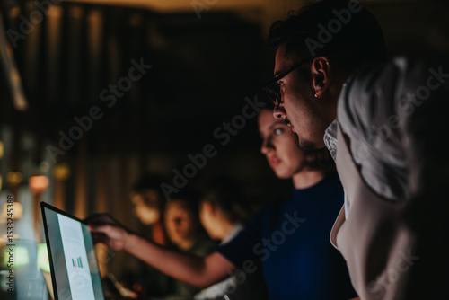 A diverse group of business people collaborate on a project, analyzing data on a computer screen during a late-night work session, striving to meet deadlines and generate innovative solutions.