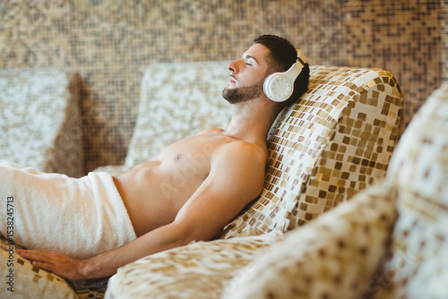 Male adult reclining on mosaic lounge chair in spa lounge, wearing white towel and headphones