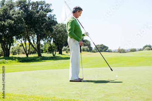 Senior golfer holding flagstick and putter on manicured green, with ball near hole