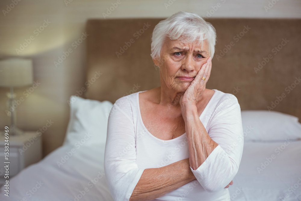 Senior woman sitting on bed in bedroom pressing hand against cheek beside nightstand lamp Senior woman sitting on bed in bedroom pressing hand against cheek beside nightstand lamp