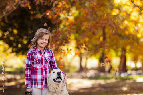 White child standing beside golden retriever in sunlit autumn park holding black leash petting dog