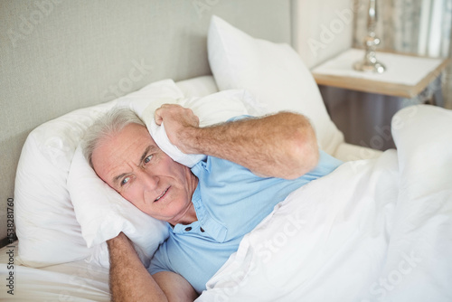 Senior man holding pillow against ear while lying in bedroom wearing light blue polo under duvet