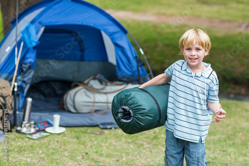 Boy carrying rolled sleeping bag while standing on grassy campsite by blue dome tent, copy space