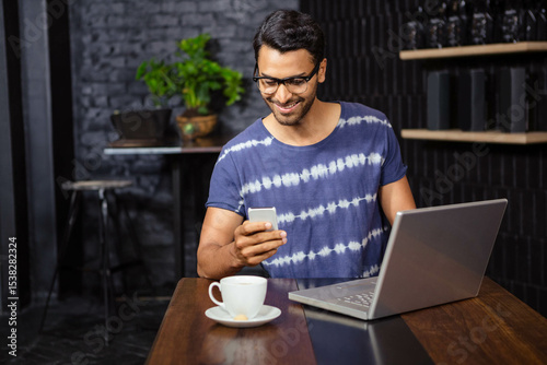 man is sitting at wooden table in café interior, holding smartphone beside laptop and coffee cup