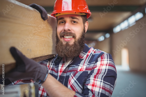 Worker carrying wooden beam on shoulder inside unfinished building, with red hard hat and gloves