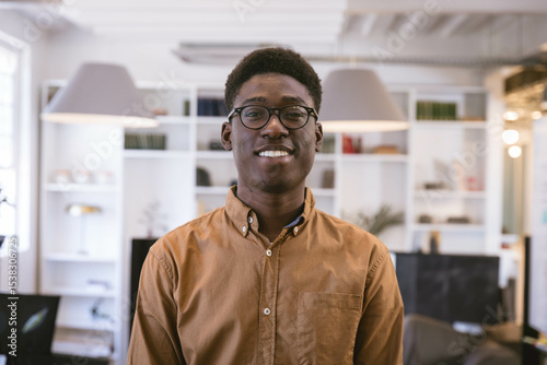 African American man standing in office wearing brown shirt, round eyeglasses against white shelves