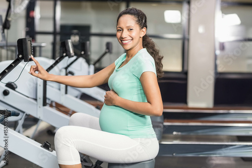 Mid adult pregnant woman wearing mint green top using rowing machine display console at gym