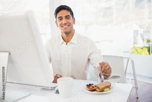 Indian man smiling and holding fork poised over sandwich and salad at office desk with monitor