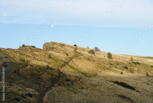 A scenic view of Arthur’s Seat, an ancient extinct volcano and the highest peak in Edinburgh’s Holyrood Park, under a baby blue sky with soft white clouds, capturing the serene Scottish landscape.