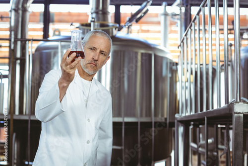 Man in white lab coat holding sample flask inside brewery with tanks, pipes, valves and platform
