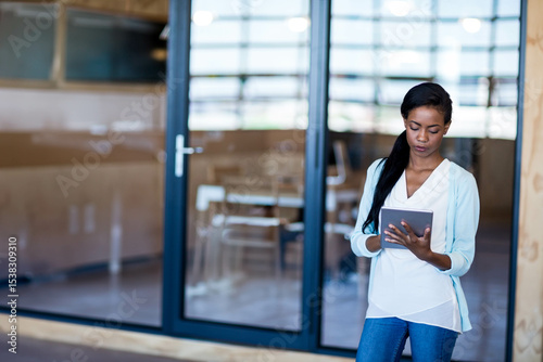 Business woman standing outside glass-walled meeting room holding tablet and viewing wooden tables