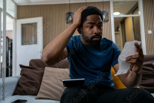 Worried young African American man looking at bills at home living room, stressed and surprised by expenses, financial concern concept
