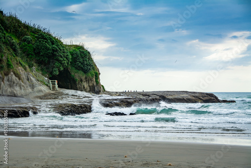 Sunset at Muriwai beach with waves splashing against the rocks and fishermen silhouettes in the distance. Auckland, New Zealand
