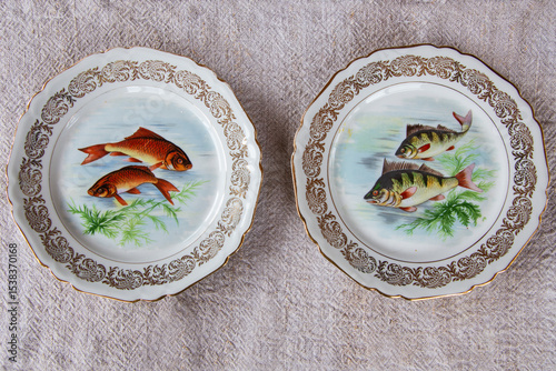 High angle view of two vintage oval porcelain dinner plates with painted fish and scalloped rim set on linen tablecloth