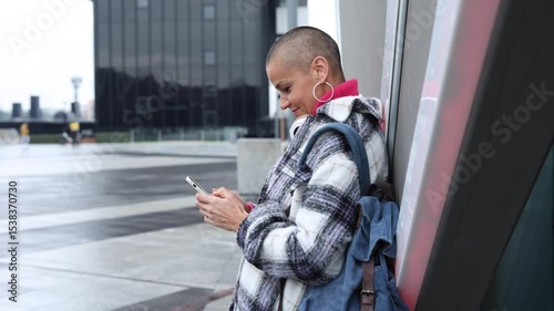 Modern urban lifestyle portrait of a stylish bald woman with a backpack leaning against a wall in the city, using her smartphone and smiling