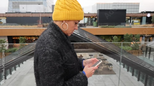 Stylish female professional wearing yellow beanie and gray coat navigating digital world through smartphone while moving across contemporary urban landscape