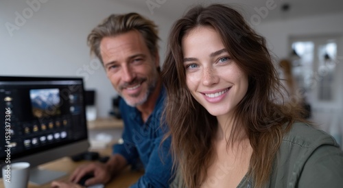 man and woman sit together at a desk filled with computer equipment, both smiling at the camera while collaborating in a bright, modern office environment during the day