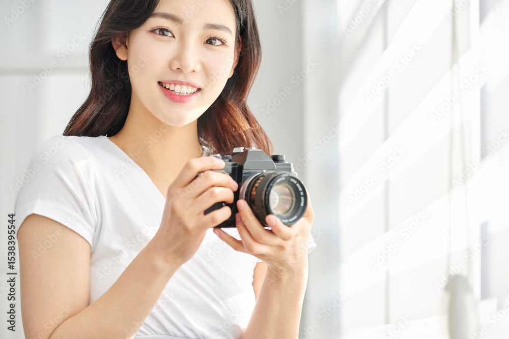 Fototapeta premium Young Asian Woman Photographer in White T-Shirt Holding Vintage Camera by Bright Window Indoors
