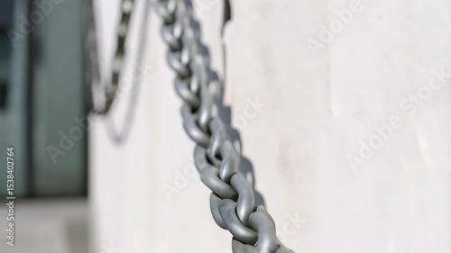 A metal chain with links hangs against a bright concrete wall casting shadow patterns in a selective focus composition on a sunny day.