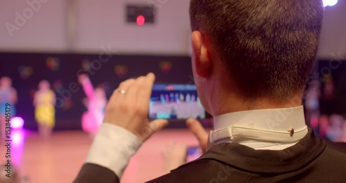 An individual is capturing a vibrant performance using their smartphone, attentively observing the dancers dressed in colorful, eyecatching outfits during a lively event they are attending
