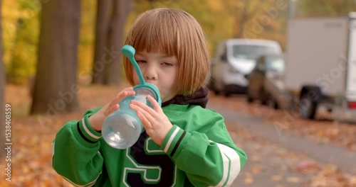 A delighted and carefree child happily sips from a bright blue bottle while joyfully strolling through a picturesque park that is beautifully adorned with vibrant autumn foliage all around